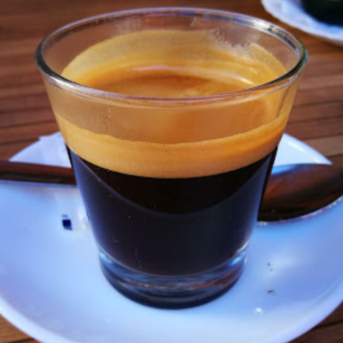 Un vaso de vidrio pequeño con un espresso con una capa espumosa en la parte superior, colocado sobre un platillo blanco con una cuchara.