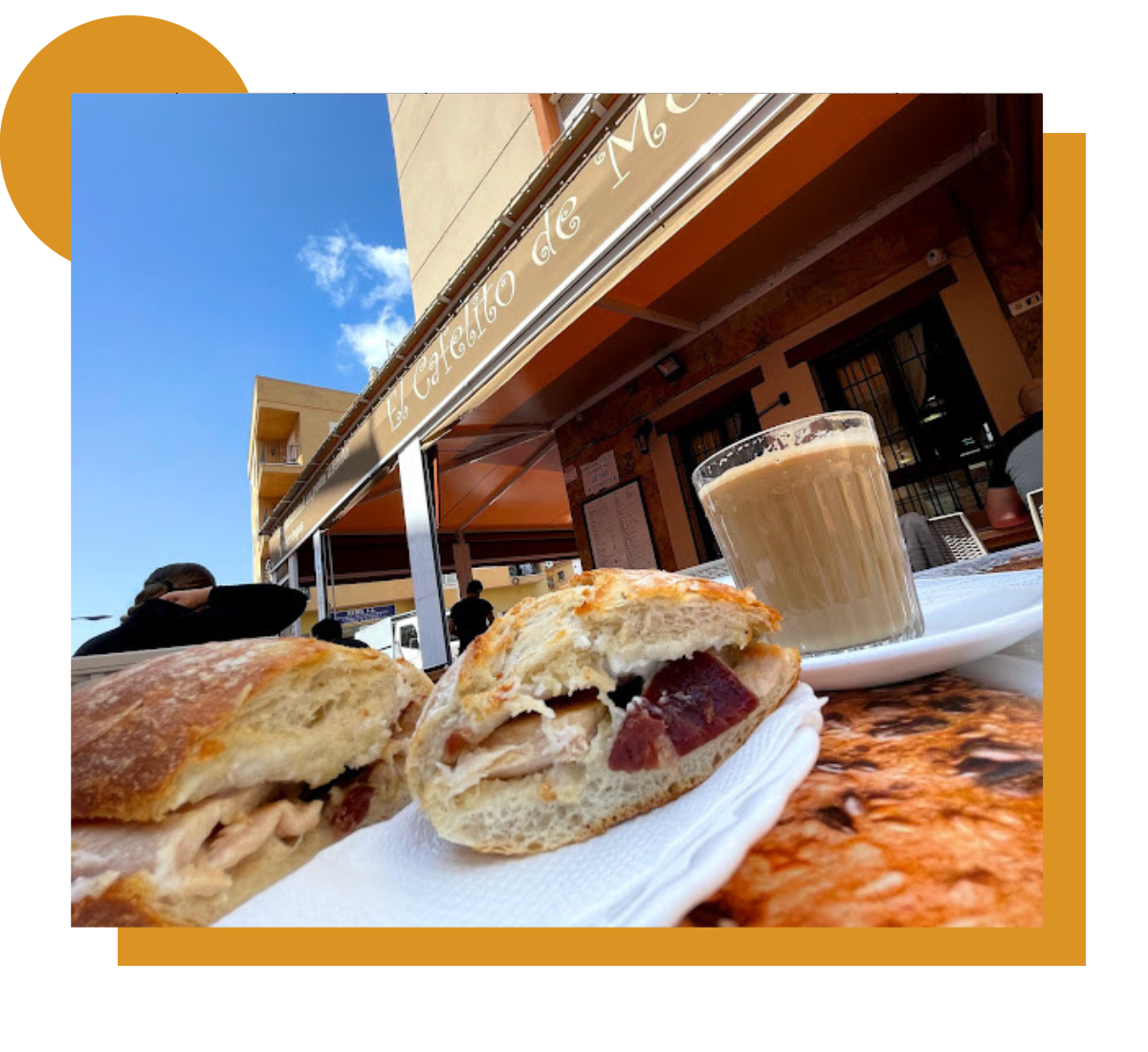 Un bocadillo con relleno de jamón y queso, servido en una mesa al aire libre junto a un vaso de café con leche. Al fondo se ve la fachada de "El Cafelito de Moy".